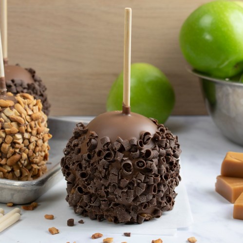 A chocolate-covered caramel apple coated in chocolate shavings, with green apples in the background.