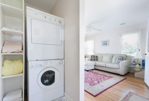 A laundry area with a stacked washer and dryer next to a bright living room featuring a sofa and decorative rug.