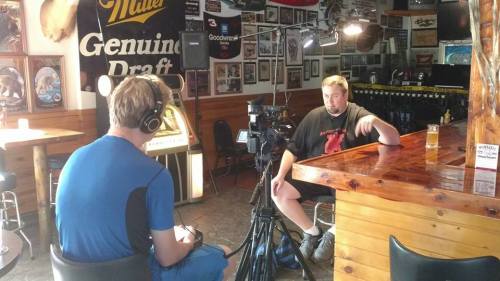 A man is being interviewed in a bar, sitting at a wooden counter with a drink, while a cameraman records.