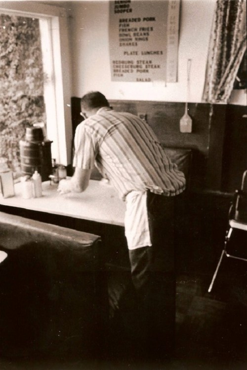 A black-and-white photo of a man in a striped shirt and apron, cleaning a table in a diner.