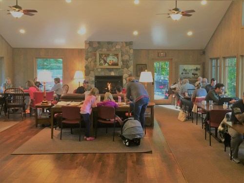 A cozy lodge interior with people socializing, a fireplace, and large windows, creating a warm, inviting atmosphere.