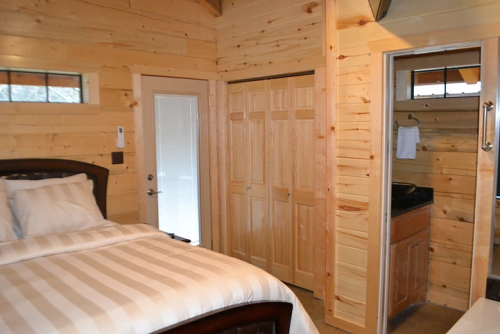 Cozy cabin interior featuring a bed, wooden walls, a closet, and a bathroom entrance. Natural light from the door.