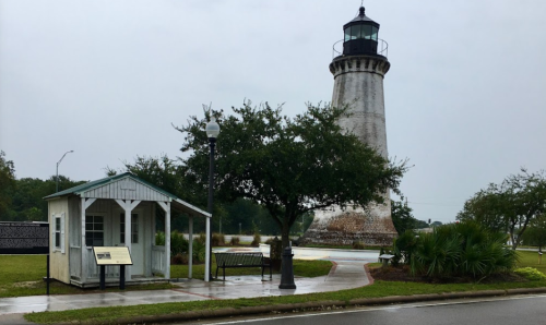 A lighthouse stands tall beside a small building and benches, surrounded by greenery on a cloudy day.