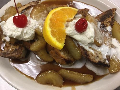 French toast topped with warm apple slices, whipped cream, cherries, and an orange slice, drizzled with syrup.