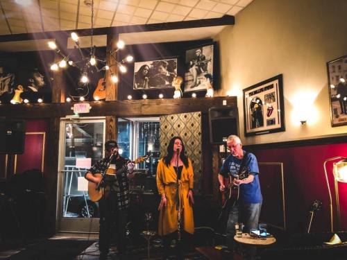 A live music performance in a cozy venue with two guitarists and a singer on stage, surrounded by framed photos.