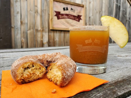 A cinnamon sugar donut and a glass of cider on a wooden table, with a rustic backdrop featuring a Yankee Cider Co sign.
