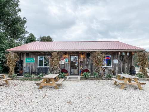 A rustic wooden building with a red roof, surrounded by picnic tables and autumn decorations, under a cloudy sky.