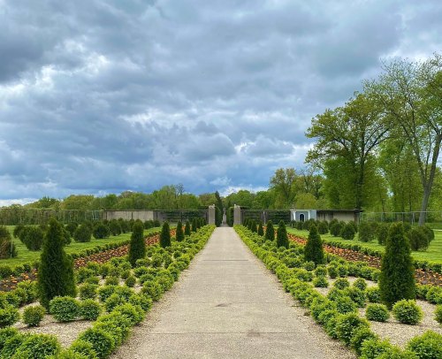 A landscaped garden path lined with shrubs and flowers, under a cloudy sky with trees in the background.
