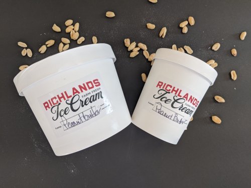Two white ice cream tubs labeled "Peanut Butter" with scattered peanuts around them on a dark surface.