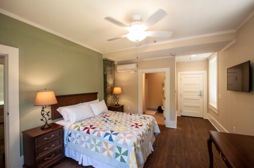 Cozy bedroom with a quilted bedspread, lamps, and a ceiling fan, featuring a door to a bathroom in the background.