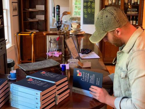 A person in a cap examines a book at a wooden counter, surrounded by stacked books and a cozy interior.