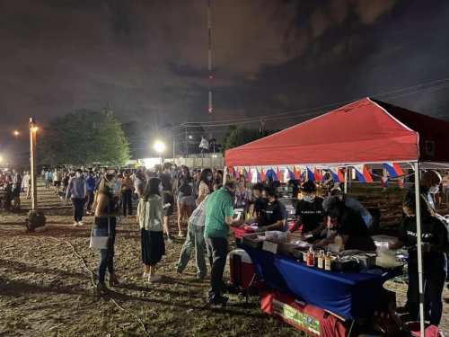A lively outdoor event at night with people gathered around food stalls under a red tent.