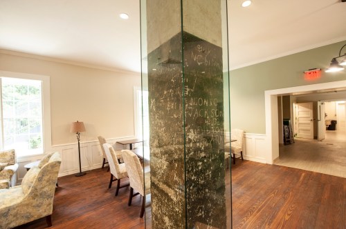 A cozy interior space with seating, a glass column, and a wall featuring faint inscriptions. Natural light from windows.