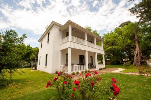 A white two-story house with a balcony, surrounded by greenery and blooming red roses in the foreground.