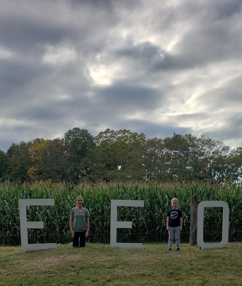 Two children stand between large letters spelling "E E O" in front of a cornfield under a cloudy sky.