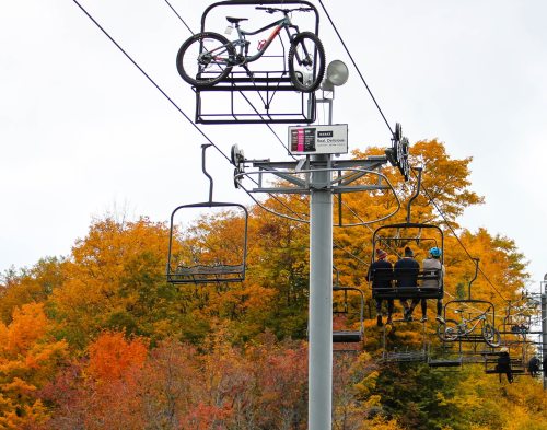 A bike on a ski lift above two people riding in a chairlift, surrounded by vibrant autumn foliage.