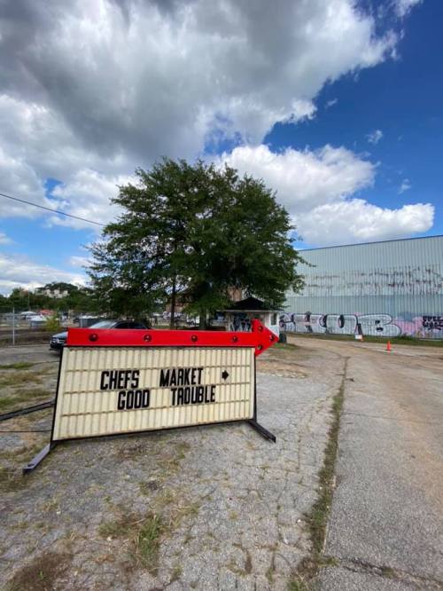 A large sign with an arrow points towards "Chef's Good Market" and "Trouble," set against a cloudy sky and graffiti wall.