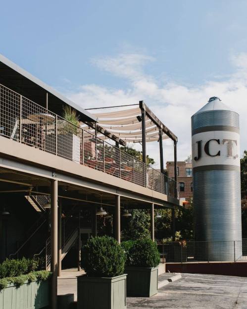 A modern building with a rooftop patio and a large silos labeled "JCT" in a sunny outdoor setting.