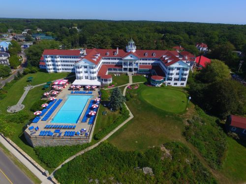 Aerial view of a large hotel with a pool, surrounded by green trees and a clear blue sky.