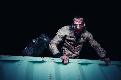 A bloodied figure in tattered clothing climbs over a container, set against a dark background.