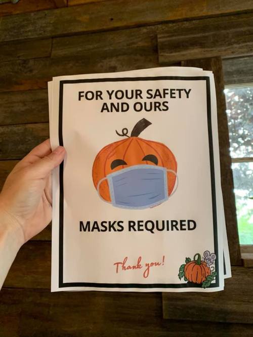 A hand holds a sign with a pumpkin wearing a mask, stating "Masks Required" for safety.