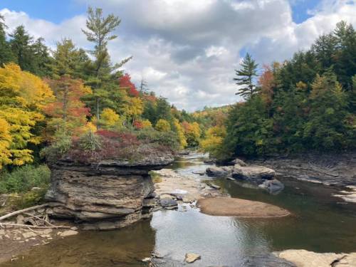 A serene river flows through a colorful forest with vibrant autumn foliage and rocky formations under a cloudy sky.