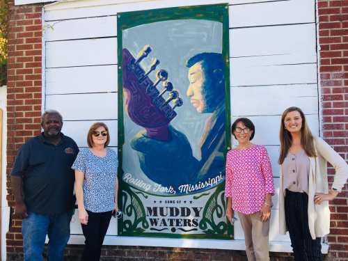 Four people stand in front of a mural of Muddy Waters in Rolling Fork, Mississippi, smiling and posing for the photo.