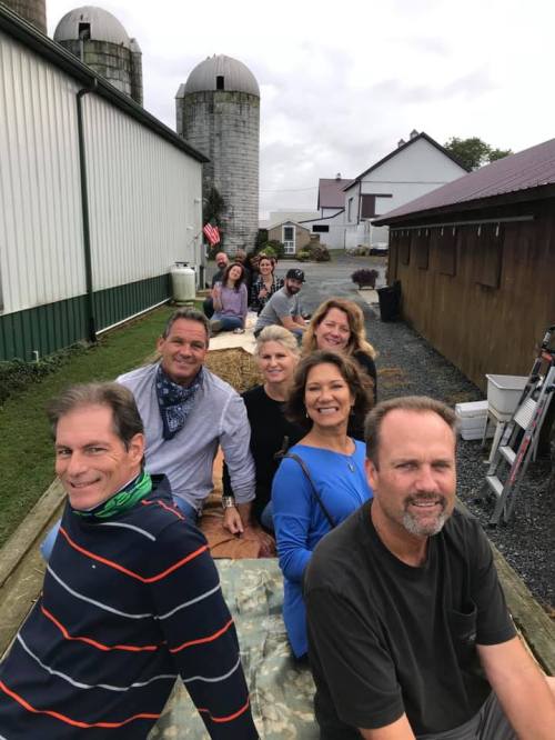 A group of people sitting on a wooden platform outdoors, with barns and silos in the background.