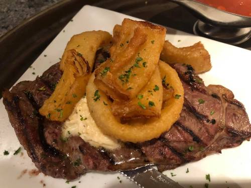 A grilled steak topped with crispy onion rings and fried potato strips, garnished with parsley on a white plate.