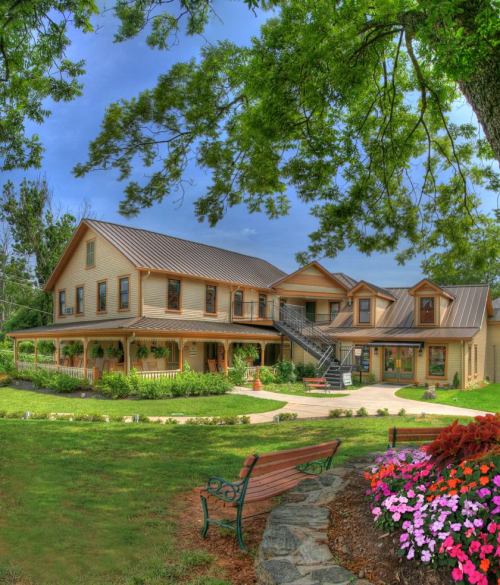 A charming two-story house with a metal roof, surrounded by greenery and colorful flowers, featuring a bench in the foreground.