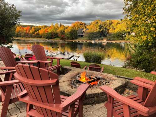 A cozy fire pit surrounded by wooden chairs, overlooking a lake with autumn foliage and cloudy skies.