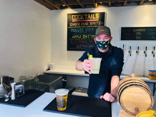A bartender wearing a mask holds a cocktail in a bright bar with a menu board in the background.