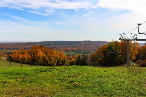 A scenic view of autumn trees with vibrant orange and yellow leaves, overlooking a green field and ski lift.