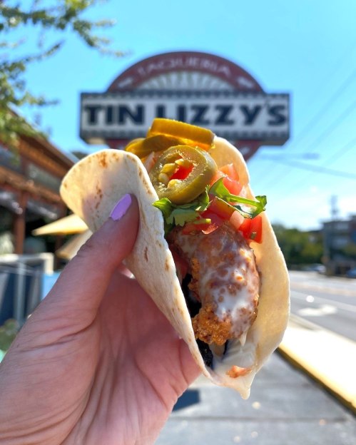 A hand holding a taco filled with toppings, with a sign for Tin Lizzy's in the background.