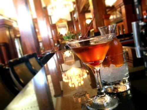 A cocktail glass with a cherry sits on a polished bar, with a carafe and glass of ice in the background.