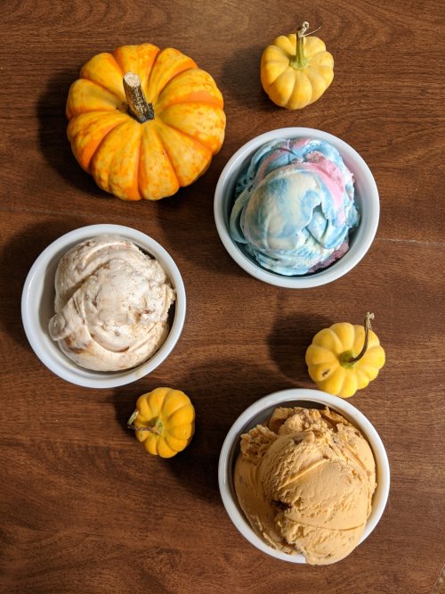 Three bowls of ice cream in various flavors, surrounded by small decorative pumpkins on a wooden table.
