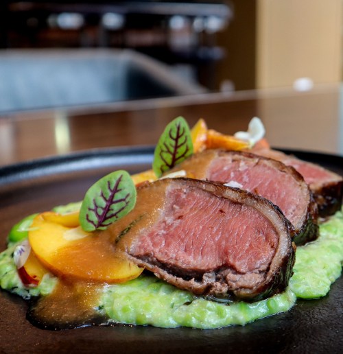 A beautifully plated dish featuring sliced beef, vibrant garnishes, and a green puree on a dark plate.