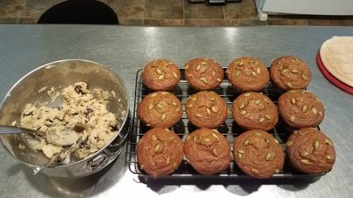 Baked muffins with pumpkin seeds cooling on a rack, next to a bowl of cookie dough on a kitchen counter.