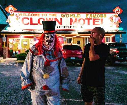 A clown in a costume stands next to a man outside the Clown Motel, with a colorful sign and dark sky in the background.