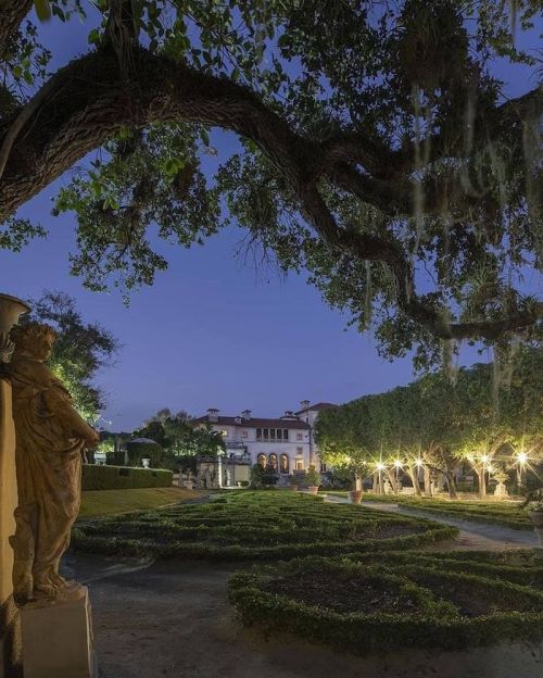 A serene garden at dusk, featuring manicured hedges, a grand mansion in the background, and softly glowing lights.
