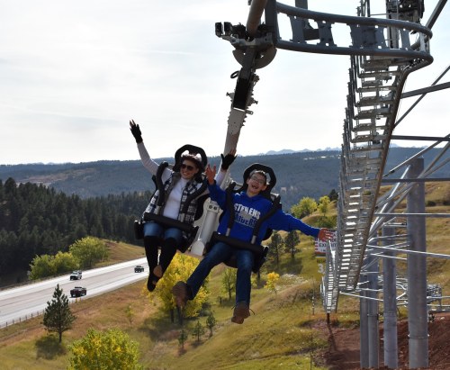 Two people joyfully riding a mountain coaster, suspended above a scenic landscape with trees and a highway below.