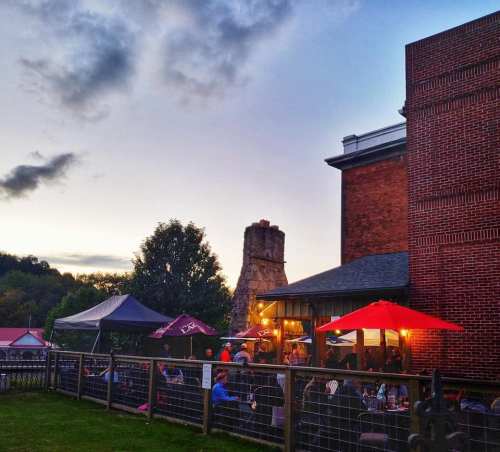 Outdoor dining area at dusk, featuring a stone chimney, red umbrellas, and a cozy atmosphere.