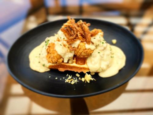 A black plate with a waffle topped with fried chicken, creamy sauce, and crispy onions, garnished with herbs.