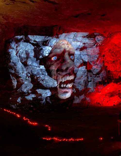 A projected face with glowing blue eyes and a grimace on a rocky wall, illuminated by red lights in a dark cave.