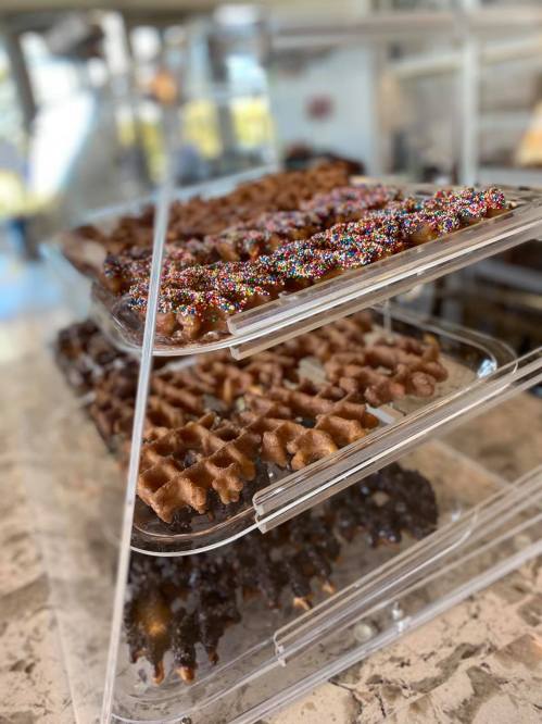 A clear display case holds stacked waffles topped with colorful sprinkles and chocolate, showcasing a variety of flavors.