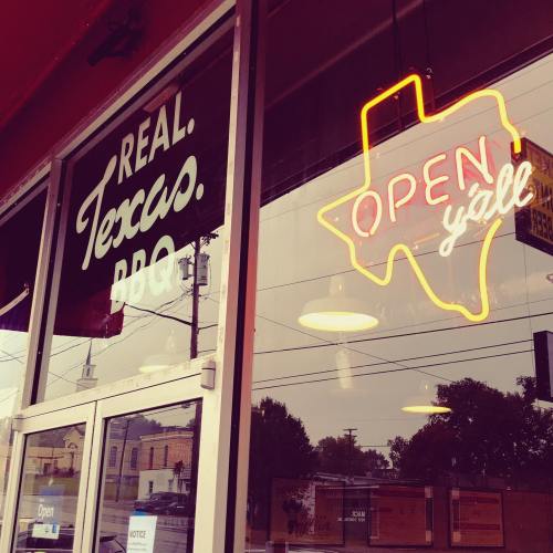A restaurant window featuring a "Real Texas BBQ" sign and a neon "OPEN y'all" sign in the shape of Texas.