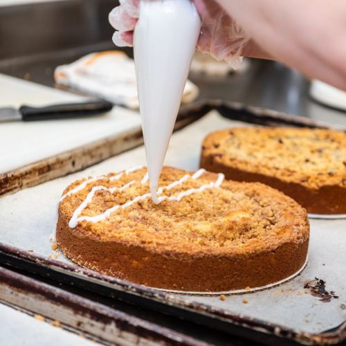 A hand decorates a cake with white icing, while two baked cakes sit on a tray in a kitchen.