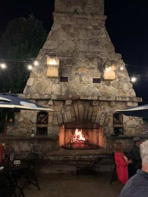 A stone fireplace with a roaring fire, surrounded by outdoor seating and string lights at night.