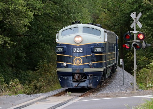 A vintage blue and yellow train numbered 722 approaches a railroad crossing surrounded by green trees.