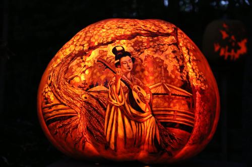 A beautifully carved pumpkin illuminated from within, depicting a woman playing a stringed instrument under a tree.